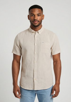 Homme portant une chemise beige à manches courtes avec poche poitrine, debout devant un fond clair uni, faisant face à l'avant.