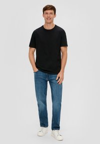 T-shirt en coton noir avec un col rond, assortie à un jean en denim bleu. Le modèle porte des baskets blanches, se tenant avec les mains dans les poches.