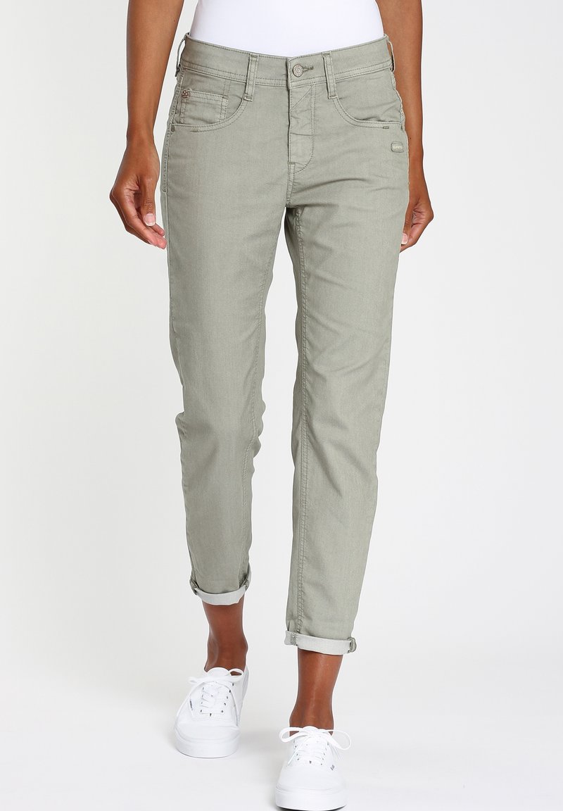 Personne portant un pantalon gris clair retroussé et des baskets blanches, debout devant un fond clair uni.