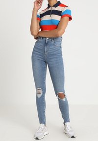 Henkilö, joka käyttää korkeavyötäröisiä revittyjä skinny-farkkuja, valkoisia sneaker-kengät ja lyhythihaista vetoketjupaitaa, jossa on punaisia, sinisiä, laivastonsinisiä ja beigejä raitoja.