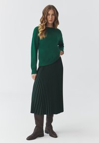 Maglione lavorato a maglia verde scuro con orlo curvo, abbinato a una gonna plissettata verde scuro. Stivali marroni fino al ginocchio completano l'outfit.