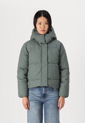 Cropped puffer jacket in muted green, featuring a high collar, stitched panels, and elastic cuffs. Paired with blue jeans.