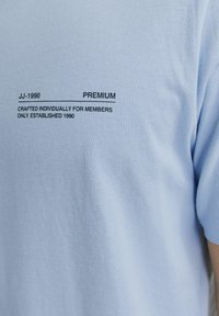 Šviesiai mėlynas medvilninis marškinėlis, su apvaliu iškirpimu ir trumpomis rankovėmis. Juodu tekstu užrašas: "JJ-1990 PREMIUM CRAFTED INDIVIDUALLY FOR MEMBERS."