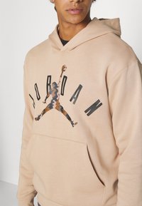 Sudadera beige con un logotipo central negro de "Jordan" y un gráfico estampado de una figura saltando. Tejido texturizado, bolsillo frontal.