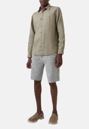 Man draagt een olijfgroen overhemd met lange mouwen en knopen, lichtgrijze shorts met textuur en beige instapschoenen, staand tegen een effen achtergrond.