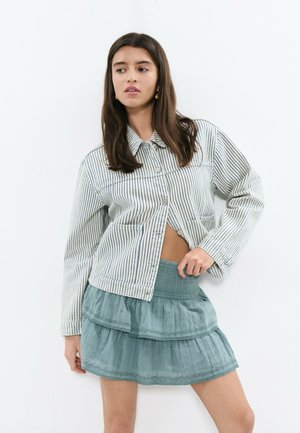 Mujer de cabello largo y oscuro, vestida con una chaqueta de rayas abotonada y una falda azul claro de volantes, posando con una mano en la cadera.