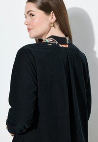Giacca di velluto a coste nera con taglio dritto, caratterizzata da una piega sul retro e un colletto a fantasia colorata. Maniche lunghe con polsini arrotolati.