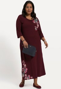 Burgundowa sukienka w kwiaty z rękawami 3/4, bocznym wzorem kwiatowym, okrągłym dekoltem oraz zwiewnym krojem, zestawiona z granatową torebką i dopasowanymi balerinami.