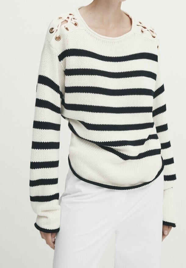 Pull en tricot blanc avec des rayures horizontales noires et des œillets dorés à lacets aux épaules, porté avec un pantalon blanc.