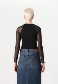 Schwarzes Langarmoberteil mit durchsichtigen Netzärmeln, kombiniert mit einem Jeansrock mit Gesäßtaschen und einem Leder-Brandlabel.