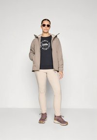 Beige vattentät jacka över en svart t-shirt med tryck, ljusbeige byxor och lila vandringskängor. Modellen bär bruna solglasögon.