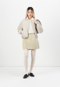 Béžová bomber bunda přes krémový pletený svetr, doplněná khaki kordovým sukněm. Černé boty a světlé punčochy dotvářejí vzhled.