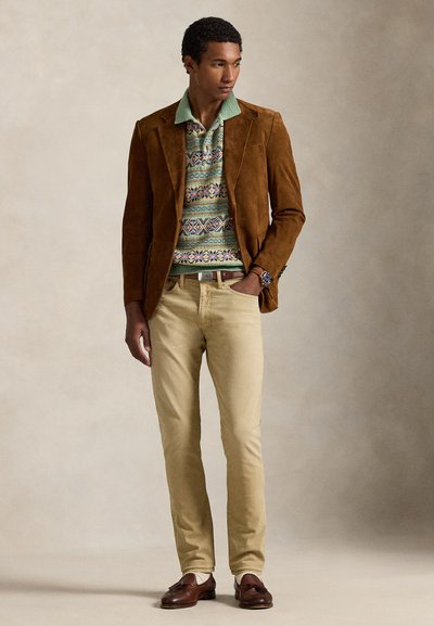 Blazer castanho em camurça sobre um suéter estampado em verde, azul e roxo, combinado com calças chino bege e mocassins castanhos.