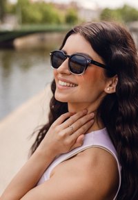 Černé obdélníkové sluneční brýle s lesklým povrchem a logem na straně, které má na sobě žena s vlnitými vlasy, v pozadí rozostřený řeky.