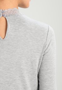 Grå långärmad topp med rund halsringning och spetsdetaljer i ryggen. Har en liten nyckelhålsöppning och knappstängning. Mjuk textur.