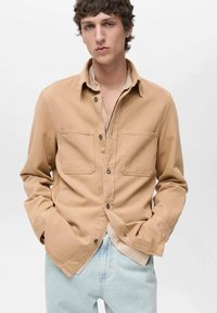 Camicia button-up tan in cotone, con due tasche sul petto, colletto e vestibilità rilassata, indossata sopra un top di colore chiaro.