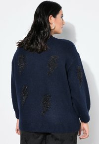 Donkerblauwe gebreide trui met zwarte appliqués in bliksemschichttextuur op de achterkant, met een losse pasvorm en geribbelde zoom.