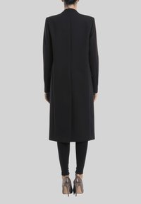 Abrigo negro largo con silueta recta, hecho de tela estructurada, sin cierres visibles y que se extiende por debajo de las rodillas.
