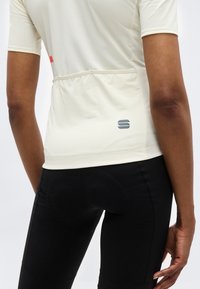 Άτομο που φοράει μπεζ κοντομάνικη φανέλα ποδηλασίας με πίσω τσέπες και μαύρο επενδεδυμένο ποδηλατικό σορτς, εμφανιζόμενο από την πίσω όψη.