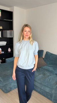 Ξανθιά γυναίκα που στέκεται σε σαλόνι φορώντας ένα ανοιχτό γαλάζιο κοντομάνικο μπλουζάκι και σκούρο μπλε παντελόνι, με το ένα χέρι στην τσέπη της.