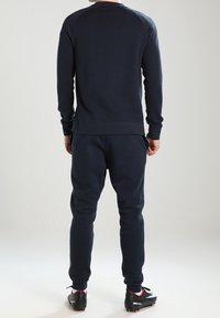 Felpa blu navy e pantaloni da jogging in tessuto morbido, con polsini a coste, vestibilità rilassata e design minimalista. Scarpe sportive nere con accenti rossi.