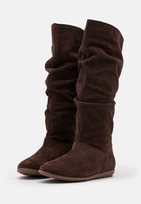 Bottes montantes en daim marron avec un design décontracté, semelles en caoutchouc plates et texture douce, présentant un bout lisse et sans pièces métalliques visibles.