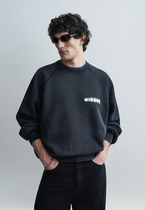 Homme portant des lunettes de soleil noires et un sweat-shirt foncé avec le logo "MISBHV", les mains dans les poches, debout devant un fond gris uni.