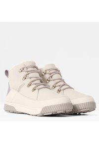 Un par de botas de invierno beiges con suelas de goma gruesa, ojales de metal, cordones beige y forro de forro polar, inclinadas contra un fondo blanco.