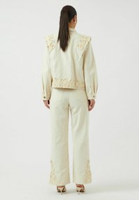 Chaqueta de mezclilla color crema con detalles florales bordados en los hombros y el dobladillo, combinada con pantalones a juego que presentan un bordado similar.