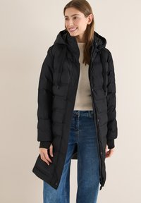 Kvinde, der smiler, iført en lang sort dunjakke med hætte, beige sweater og blå jeans, står foran en ensfarvet baggrund.