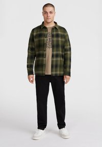 Groene geruite flanellen shirt over een beige T-shirt, gecombineerd met zwarte broek en witte sneakers. Het shirt heeft knopen en een casual pasvorm.