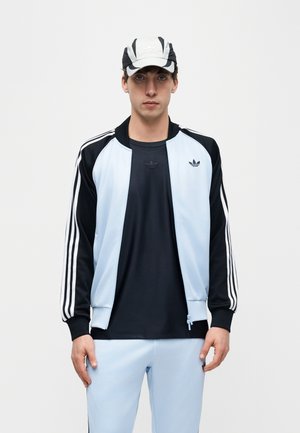 Giovane uomo che indossa una tuta Adidas azzurra e nera con strisce e un cappellino abbinato bianco e nero, in piedi davanti a uno sfondo uniforme.
