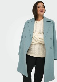 Gaisni zils vilnas mētelis ar divrindu dizainu, platām atloks un diviem priekšējiem pogām, uzvilkts virs krēmkrāsas adītā vītne džempera un melnām biksēm.