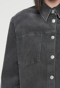 Black corduroy jacket featuring a pointed collar, front pocket, and silver snap buttons. The fabric has a textured, ribbed appearance.