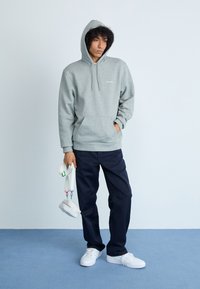 Felpa grigia con cappuccio e tasca a marsupio, abbinata a pantaloni blu e scarpe da ginnastica bianche. Gli accessori includono una borsa piccola con clip colorate.