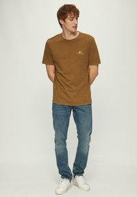 Bruine katoenen t-shirt met een ronde hals, korte mouwen en een klein tekstdetail aan de voorkant. Gecombineerd met blauwe jeans en witte sneakers.