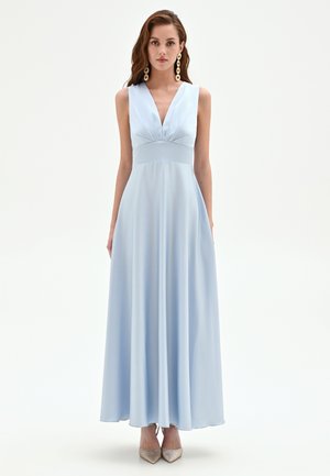 Donna che indossa un abito senza maniche, azzurro chiaro, lungo fino al pavimento con scollatura a V profonda, abbinato a orecchini dorati pendenti e tacchi a punta.