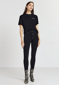 Donna in piedi, indossa una maglietta nera "Sex and the City", jeans skinny neri a vita alta e stivaletti alla caviglia con stampa serpente.