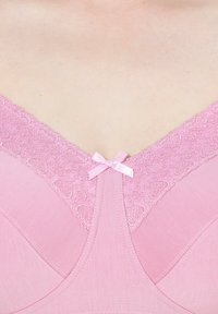 Vaaleanpunainen rintaliivi, jossa on pitsireunus sydänkuvioilla, pieni vaaleanpunainen satiininauha keskellä etupuolta ja sileä kangas saumattomalla designilla.