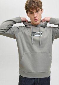Sudadera gris con bolsillo tipo canguro en el frente, capucha con cordones y un logotipo en blanco y negro en el pecho. Tejido suave y texturizado.