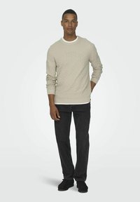 Pull en beige clair texturé avec un motif en losange, poignets et ourlet côtelés, associé à un jeans foncé et des chaussures noires. Design simple et moderne.