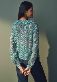 Blouse légère à manches longues avec un motif floral turquoise, en tissu transparent, encolure ronde et détails froncés aux épaules, associée à un jean foncé.