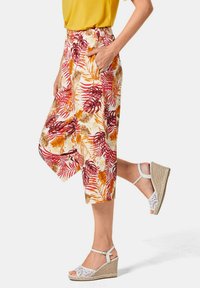 Culottes fleuris blancs, rouges et oranges avec des motifs de feuilles. Comprend des poches latérales. Associé à des sandales compensées blanches et un top jaune.