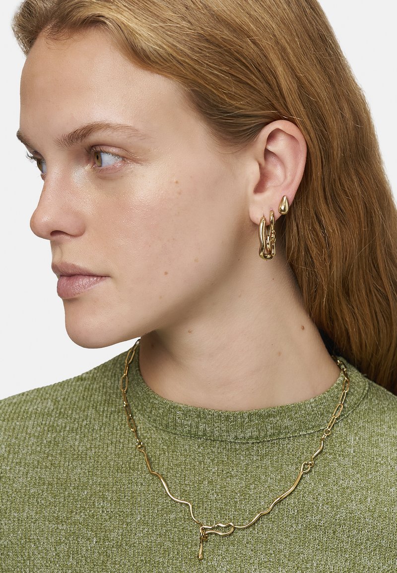 Goldene Schmuckstücke aus Metall mit unregelmäßigen Formen; hängende Creolen und eine wellenförmige Ketten-Halskette. Vor einem strukturierten grünen Pullover präsentiert.