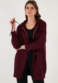 Bordo siuvinėta striukė su kapisonu, su tekstūruota medžiaga ir priekinėmis kišenėmis, dėvima ant juodos bluzės ir su juodomis kelnėmis.