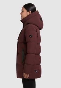 Burgundi pufi dzseki kapucnival, fekete cipzárokkal és logóval ellátott folttal a hajtókán. A design steppelt és laza szabású.