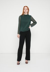 Green long-sleeve top with sheer sleeves paired with black wide-leg trousers and black heels. Simple design, smooth texture.