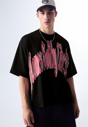 Mladý muž v černém oversize tričku s velkým červeným zkresleným textem a světle béžovou baseballovou čepicí, stojící před jednobarevným pozadím.