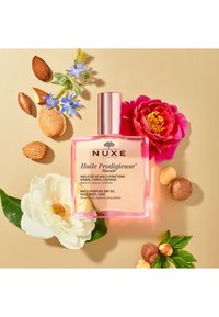 NUXE multifunktionelles Trockenöl in einer klaren rosa Flasche mit einem metallischen Deckel, umgeben von weißen Blumen, Mandeln und grünen Blättern auf einem beigen Hintergrund.