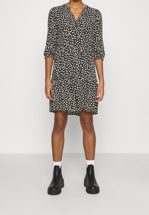 Femme portant une robe noire à imprimé floral avec des manches trois-quarts, associée à des chaussettes blanches et des bottines noires épaisses.